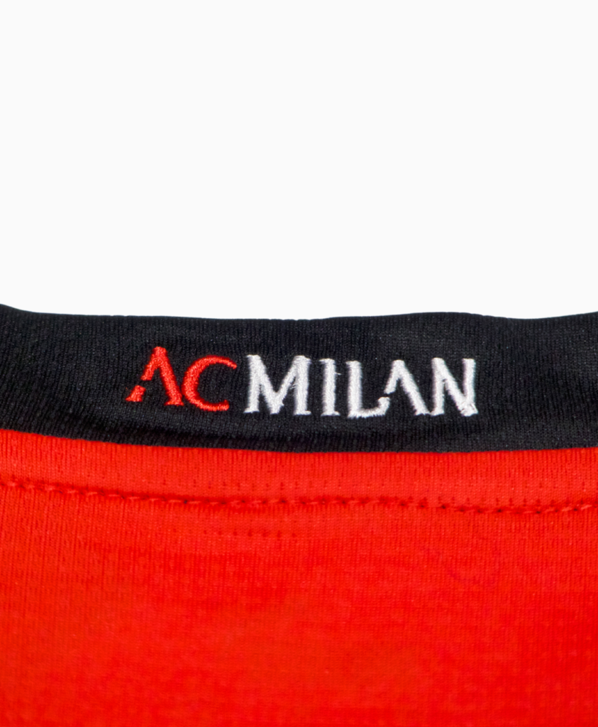 Imagen del detalle del cuello de la camiseta de fútbol del AC Milan de la temporada 2023-2024, con el texto "AC Milan" en color rojo y blanco.